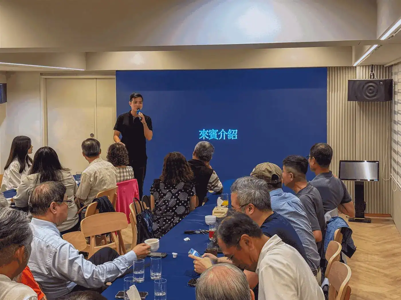 萬聚所包場例會現場，講者拿麥克風致詞介紹，社員圍長桌而坐