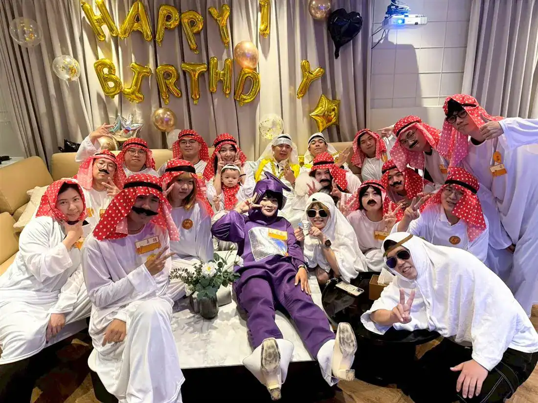 萬聚所勤美館阿拉伯主題變裝生日派對大合照：多數人戴紅底白點頭巾、穿白長袍，有人只披白毛巾，布簾上掛 HAPPY BIRTHDAY 氣球字